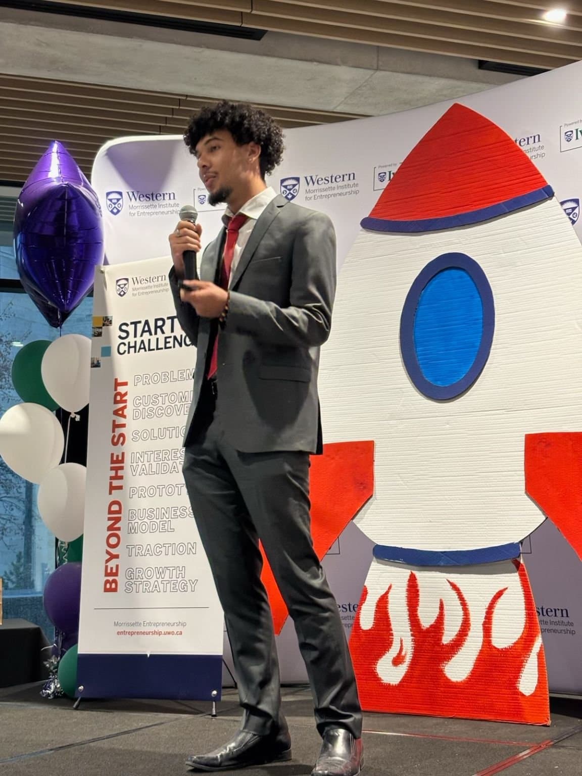 Kalan Roye pitching Crux Planner on stage at the Western Morrissette Institute Startup Challenge, with a START CHALLENGE banner and rocket cutout behind him